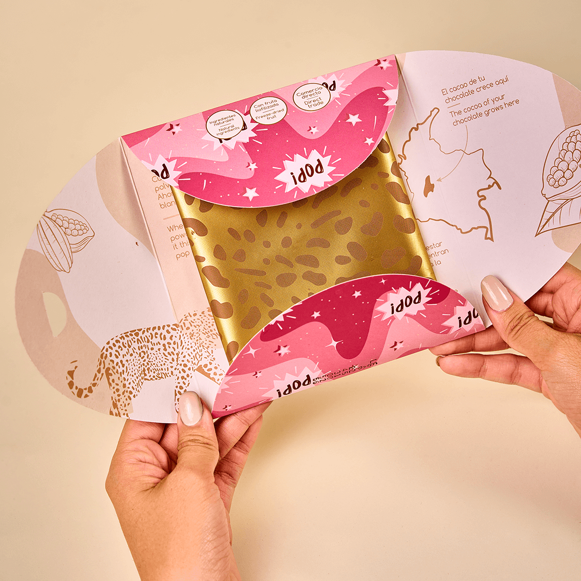 Dos manos sostienen una barra de chocolate blanco con fresa y Pop Rocks de Karü; el empaque principal está ligeramente abierto, dejando ver un poco del envoltorio secundario cerrado.