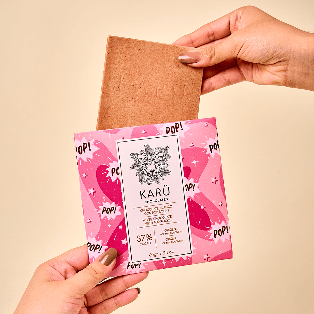 Manos sosteniendo la barra de chocolate blanco con fresas y pop rocks (“Quipitos”) de Karü, enfocando el detalle del empaque y la textura crujiente.