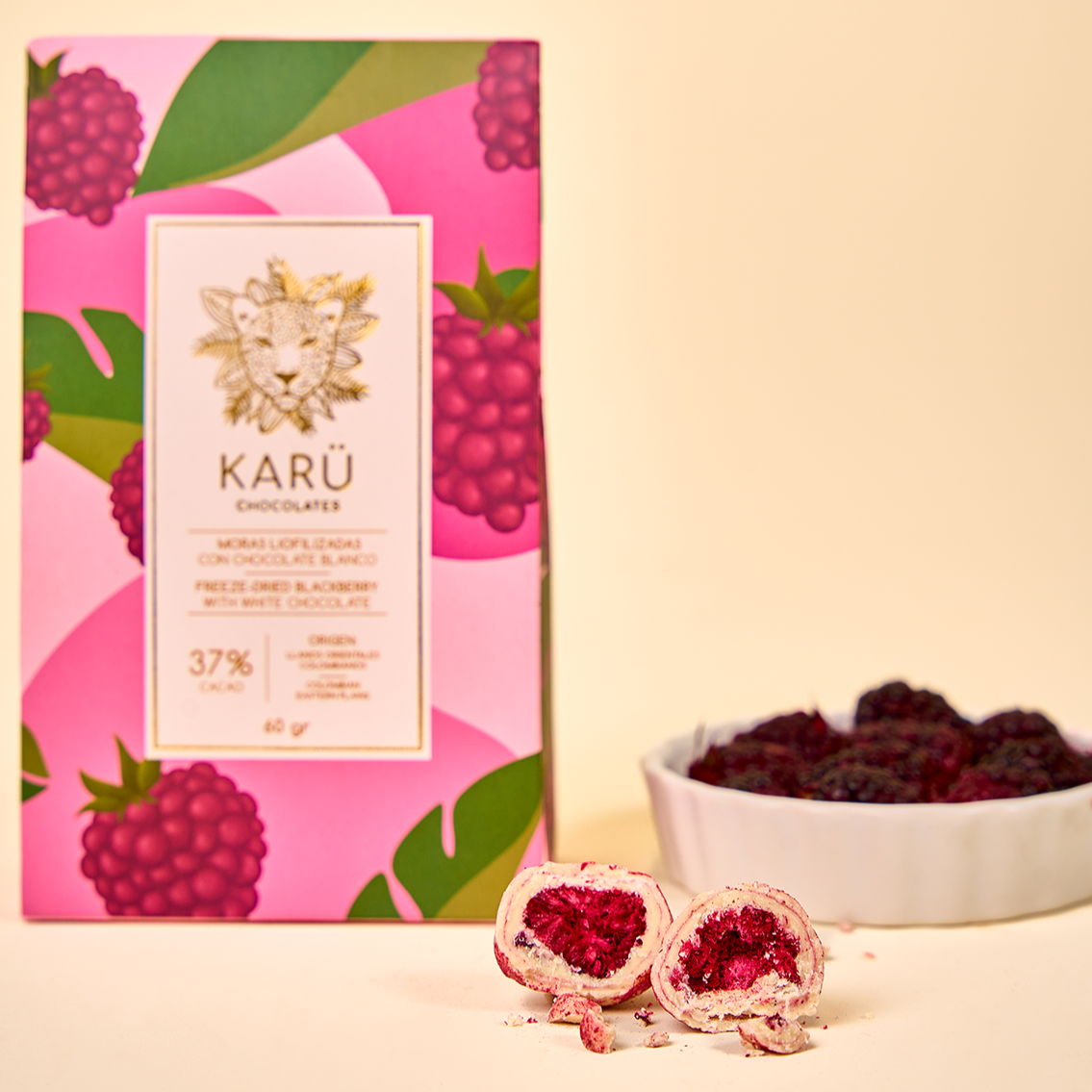Caja de moras cubiertas con chocolate blanco, dos moras partidas al frente y un recipiente con moras frescas al fondo