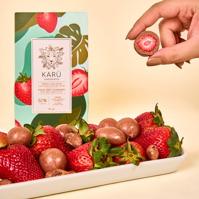 Caja de fresas liofilizadas cubiertas con chocolate de leche Karü, acompañada de una bandeja con fresas cubiertas, fresas frescas sin cobertura y una mano sosteniendo una fresa.
